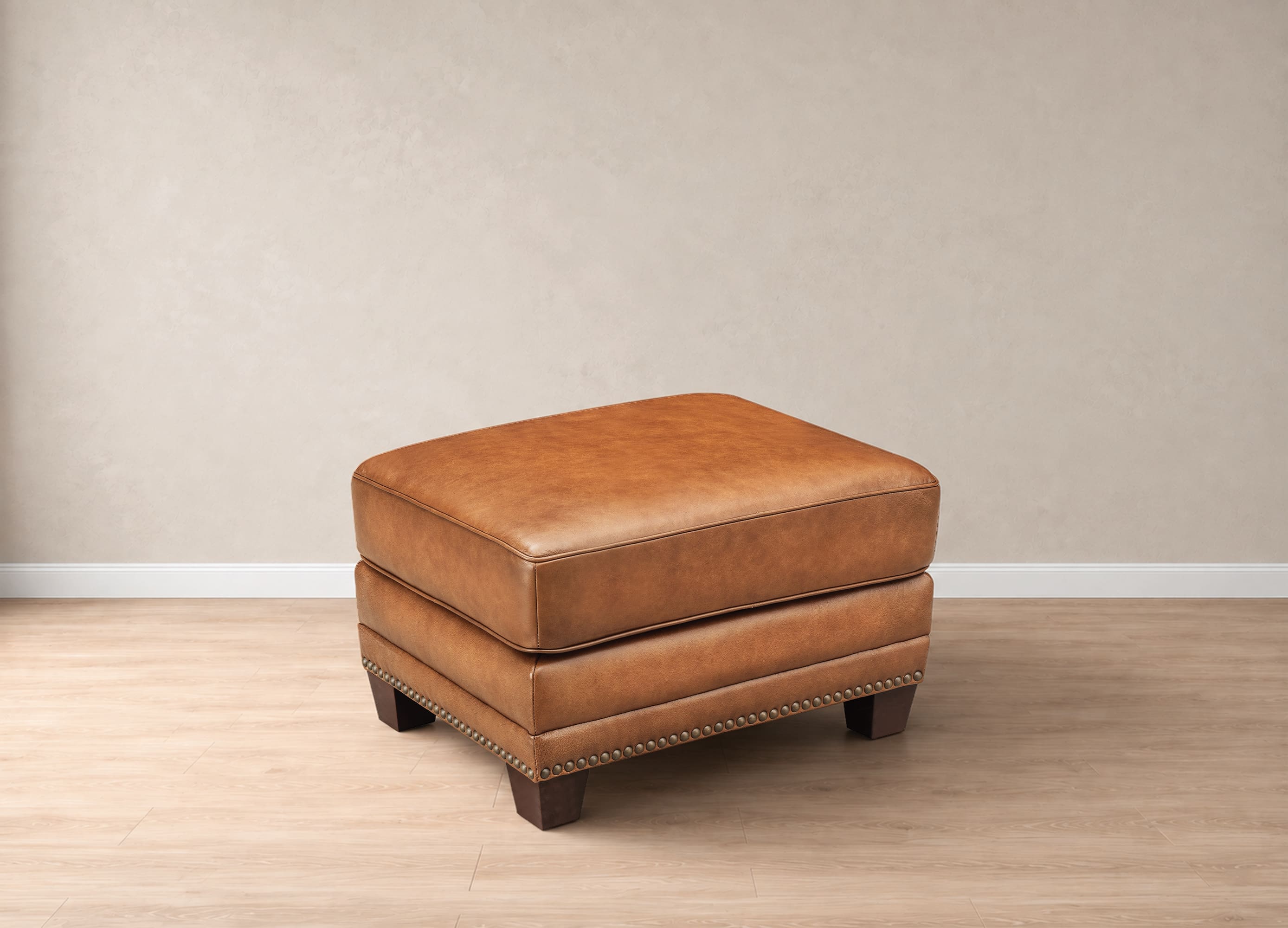 A square brown leather ottoman with dark wooden legs and nailhead trim, set against a neutral beige wall and light wood floor.