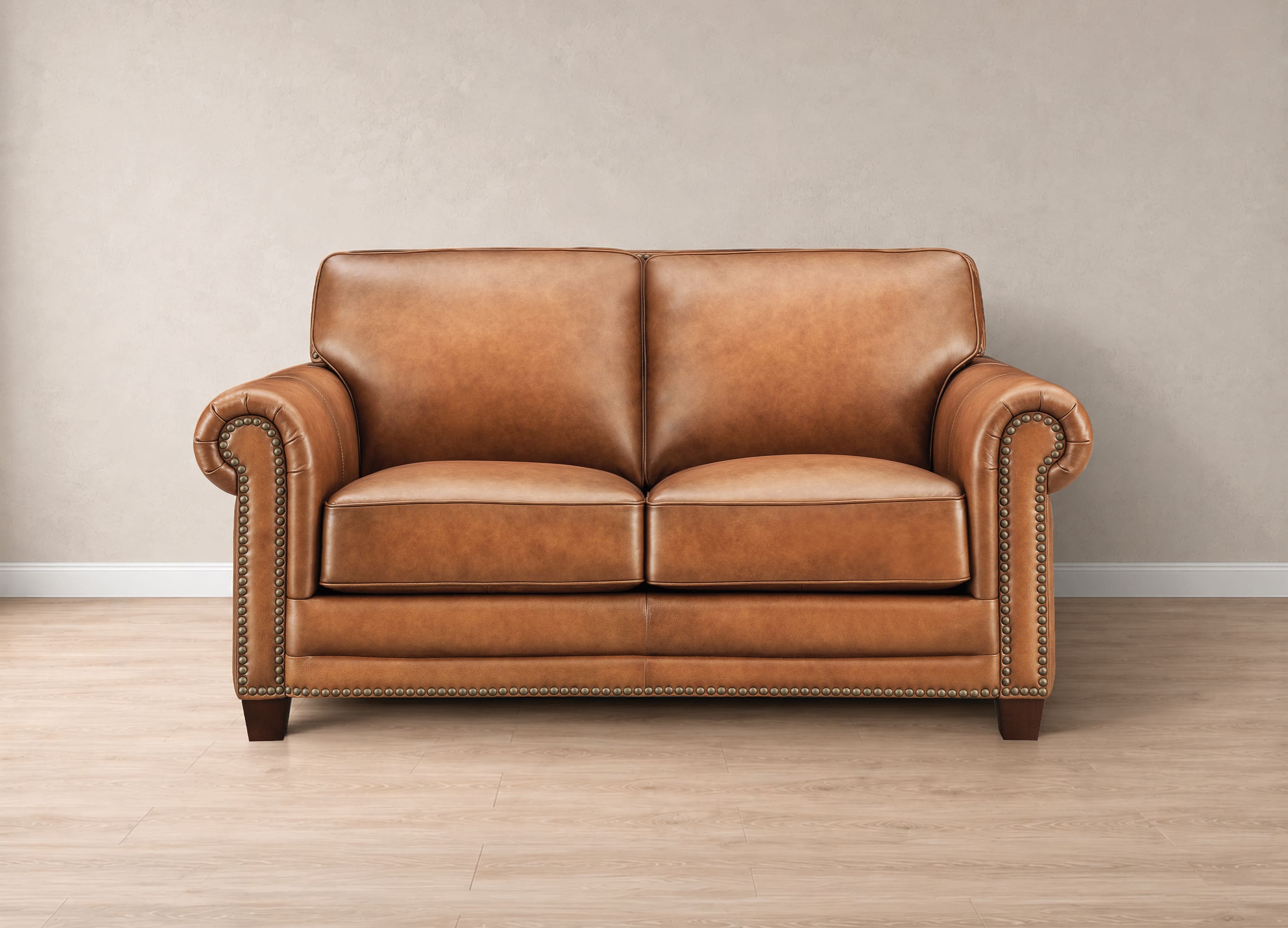 A classic two-seater loveseat sofa upholstered in warm tan leather, featuring rolled arms with nailhead trim, dark wooden legs, and a clean studio setting with a neutral beige wall and light wood floor.