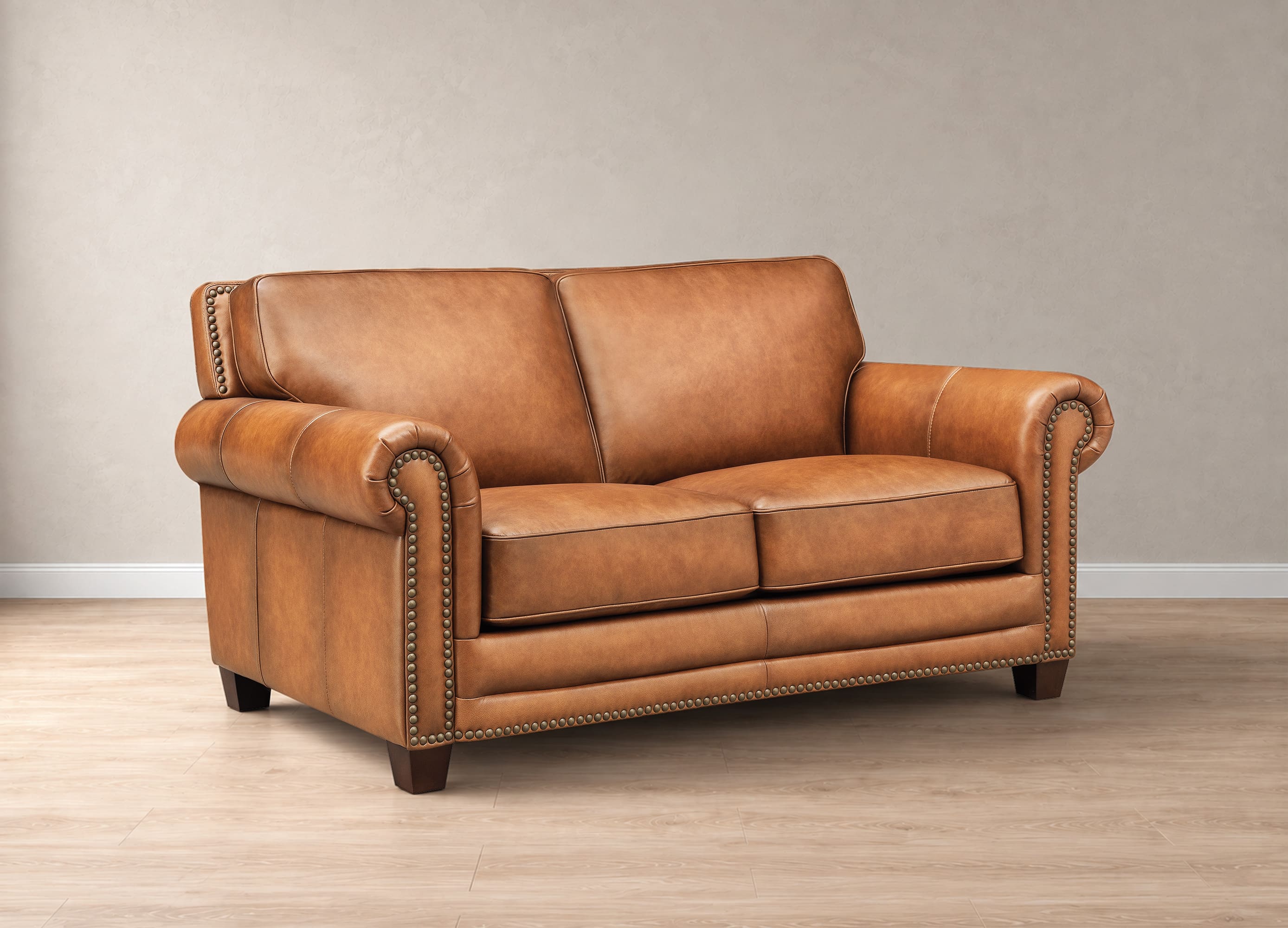 A classic two-seater loveseat sofa upholstered in warm tan leather with rolled arms, nailhead trim, and dark wooden legs, set against a neutral beige wall and light wood floor.
