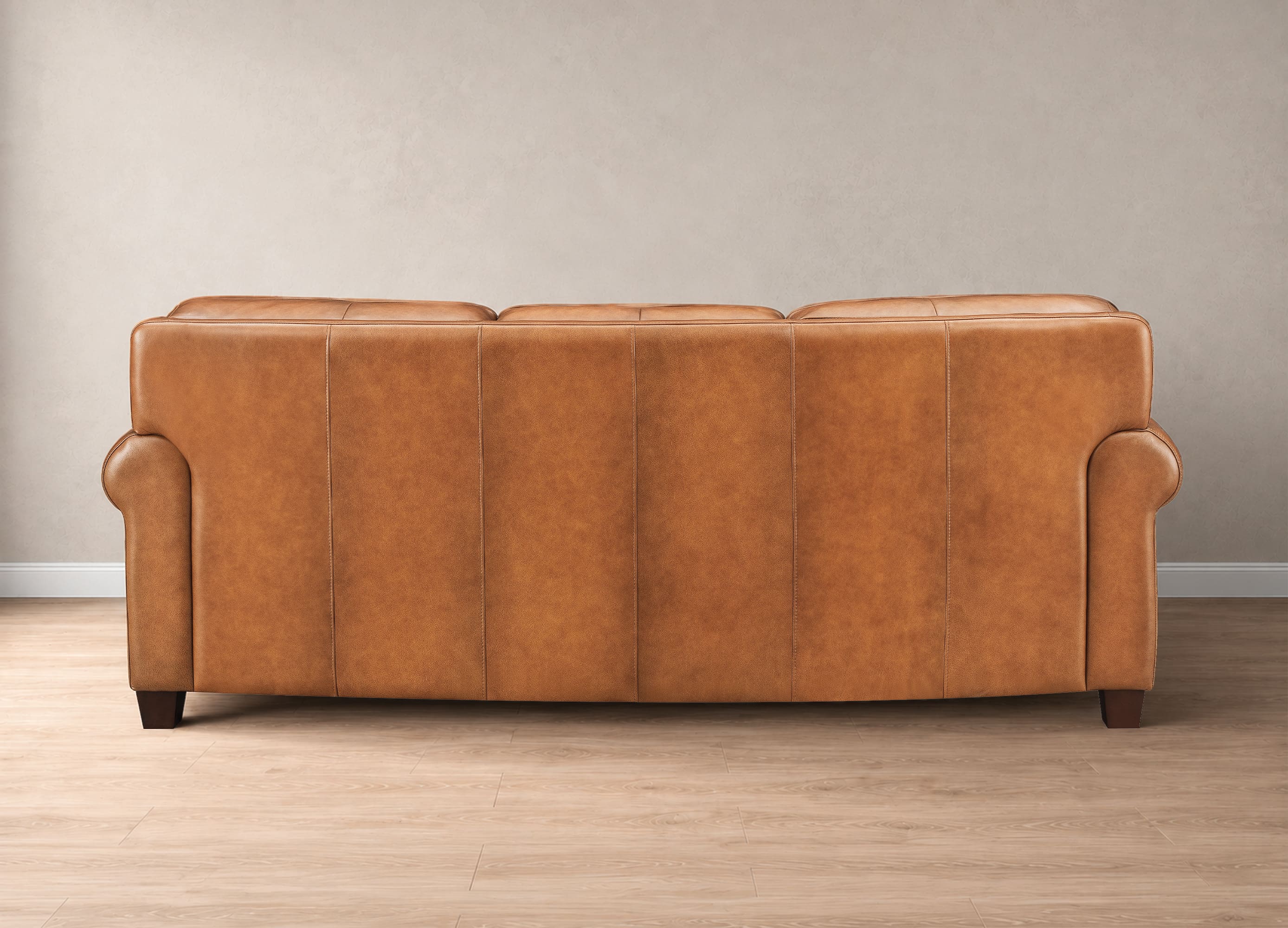 A three-seater sofa upholstered in warm tan leather, shown from the back against a neutral beige wall and light wood floor.