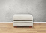 White ottoman with wooden legs on a wooden floor against a gray wall