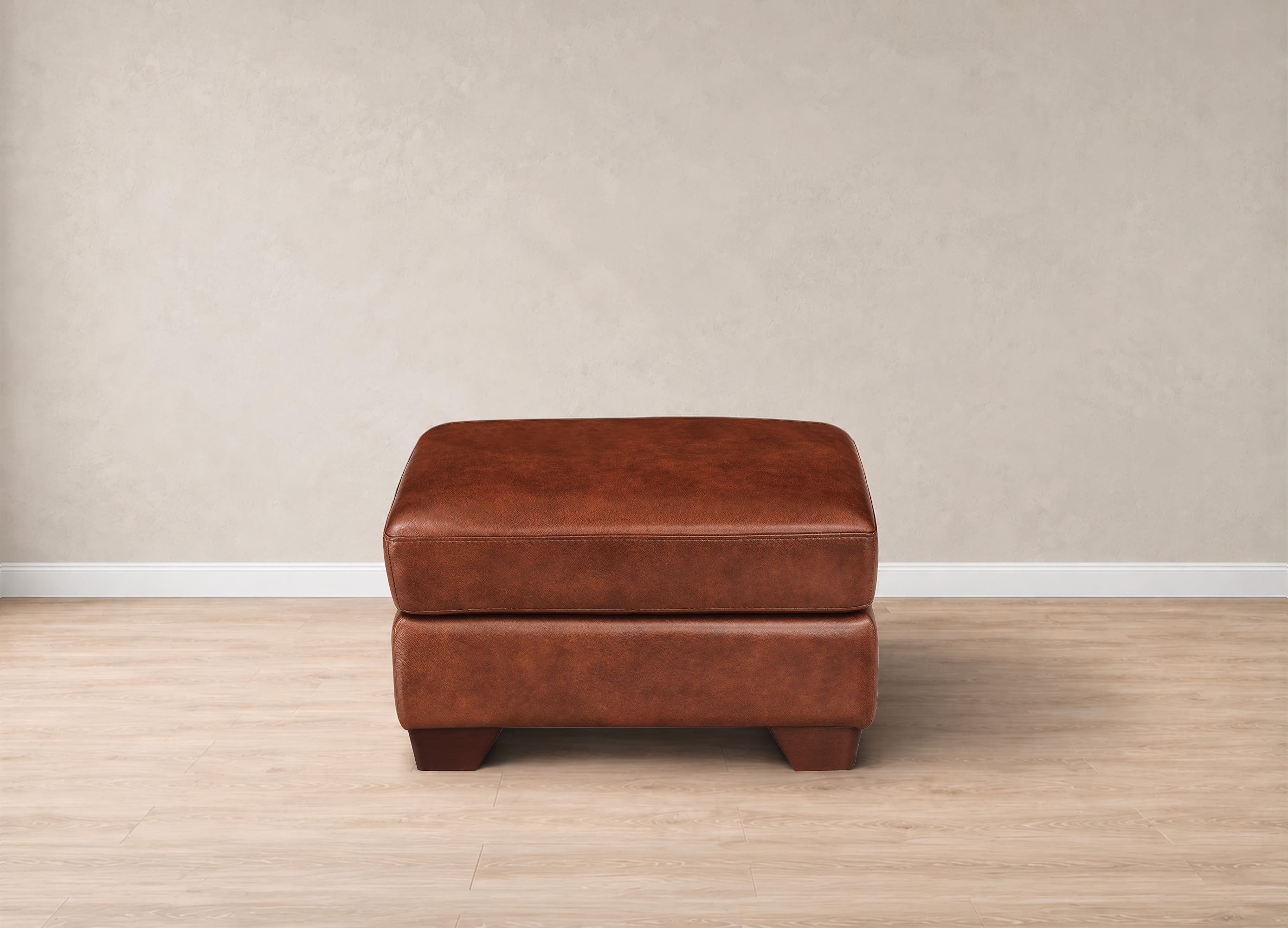 A square brown leather ottoman with a two-tiered design, standing on a light wood floor against a textured beige wall.