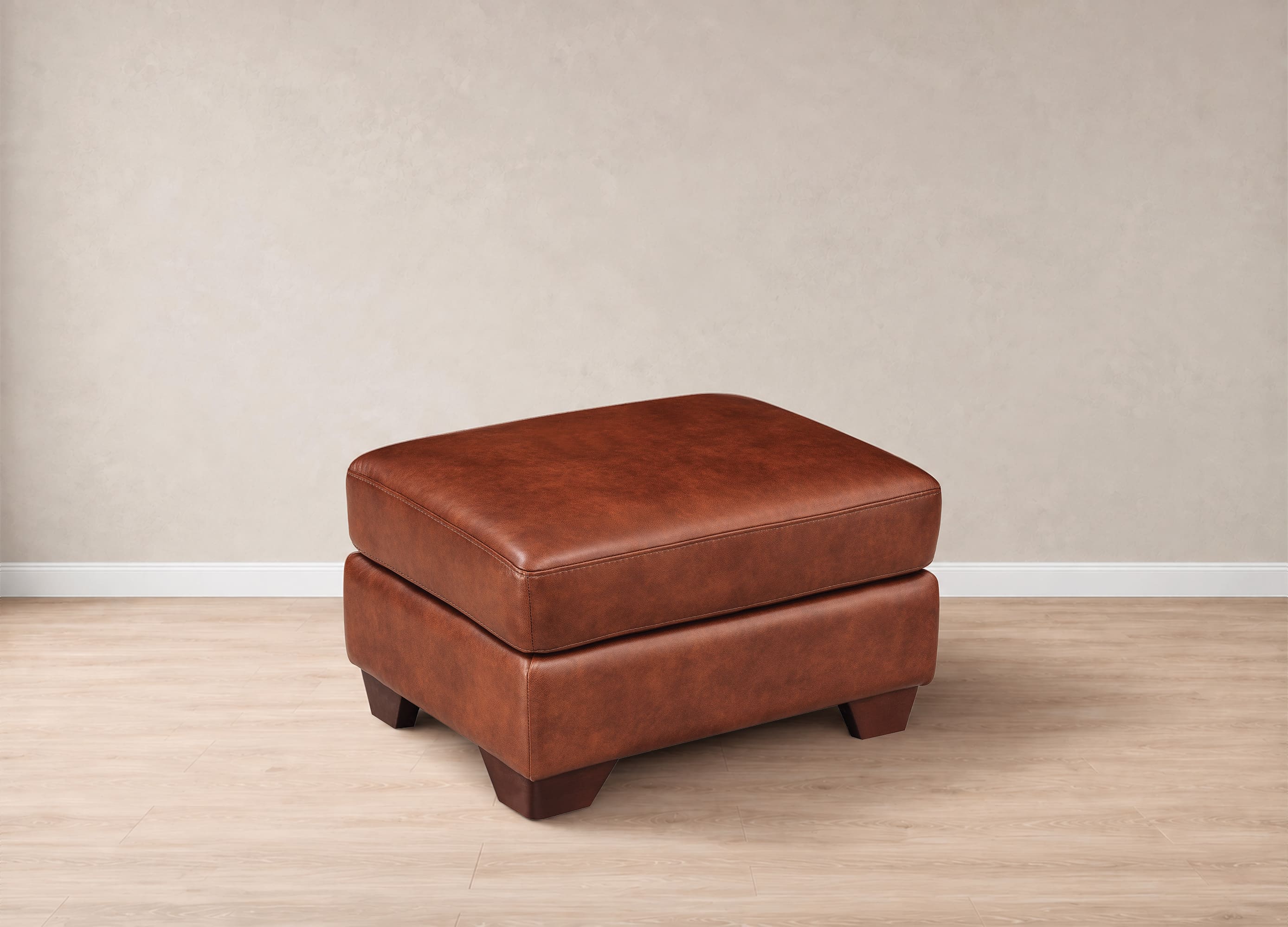 A brown leather ottoman with a stacked design and dark wooden legs, set against a neutral beige wall and light wood floor.