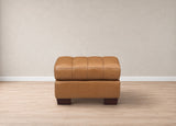 A tan leather ottoman with a tufted top and dark brown wooden legs, positioned against a neutral beige wall and light wood floor.