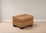 A tan leather ottoman with a tufted top cushion and dark brown block legs, set against a neutral beige wall and light wood floor.