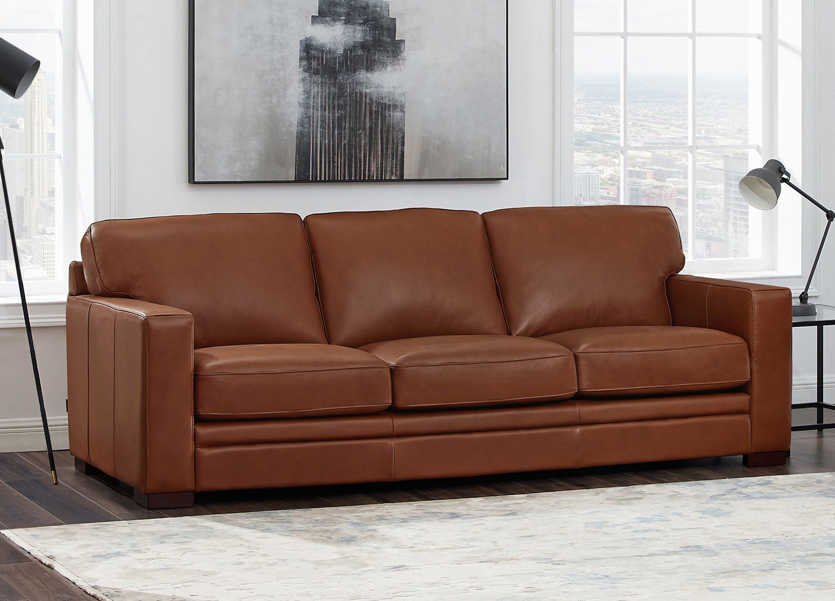 Brown leather sofa in a living room with large windows and abstract art on the wall.